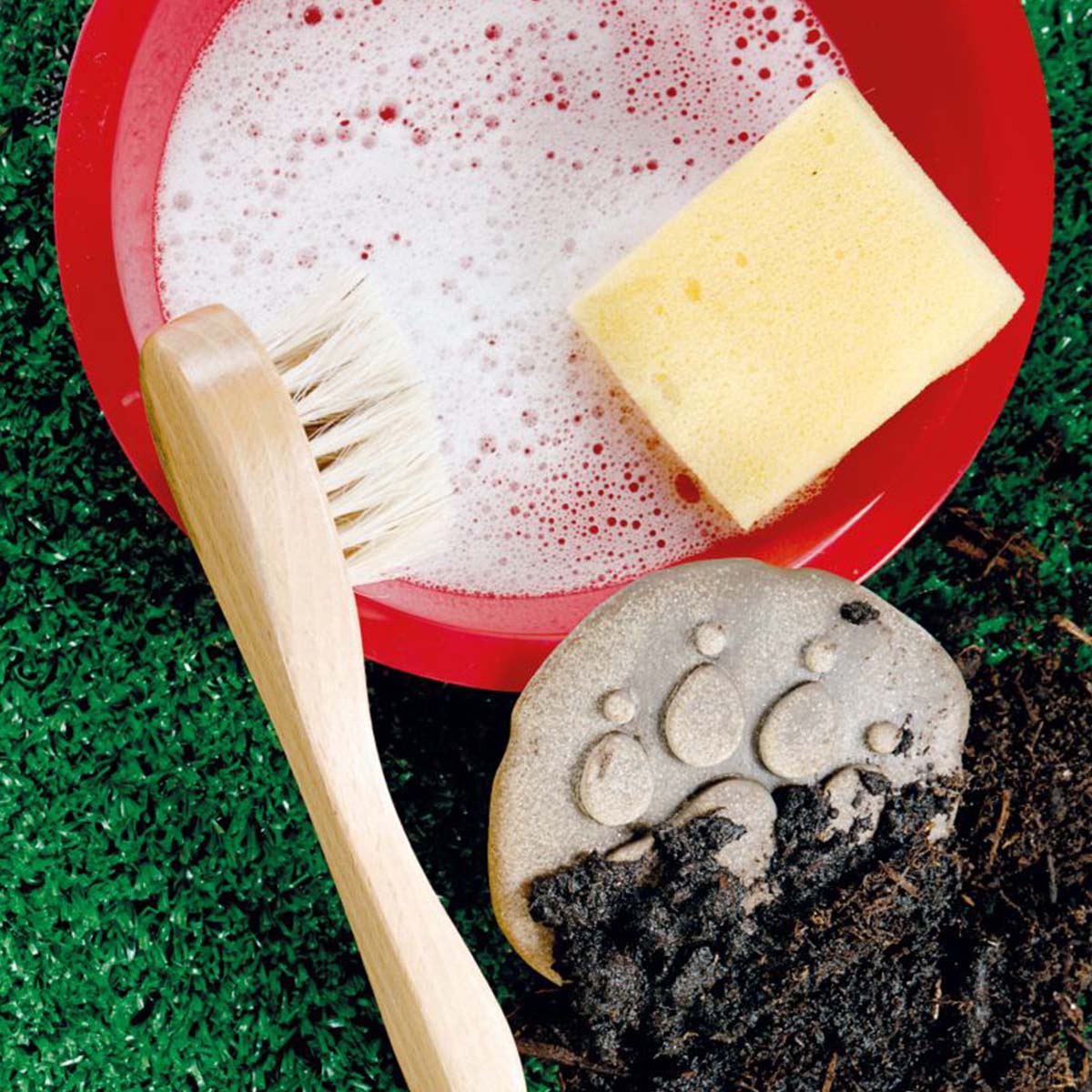 kamenná stopa psa zanesená pôdou vedľa červenej misky so saponátovou vodou hubkou a kefkou